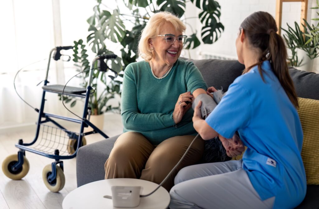 A smiling older adult sits on a couch while a young nurse is taking their blood pressure.